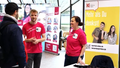 Studierender unterhält sich an einem Messestand mit Mitarbeitenden des Unternehmens.