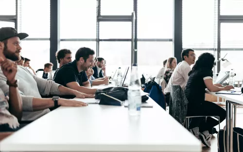 Erwachsene sitzen aufmerksam in einem hellen Seminarraum und folgen konzentriert dem Unterricht; einige arbeiten an Laptops.