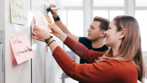 Zwei Personen arbeiten gemeinsam an einem an einem Whiteboard und heften Notizzettel daran.