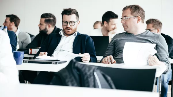 Mehrere Männer die in einem Seminarraum sitzen und dabei vertieft in Unterlagen und Gespräche wirken.